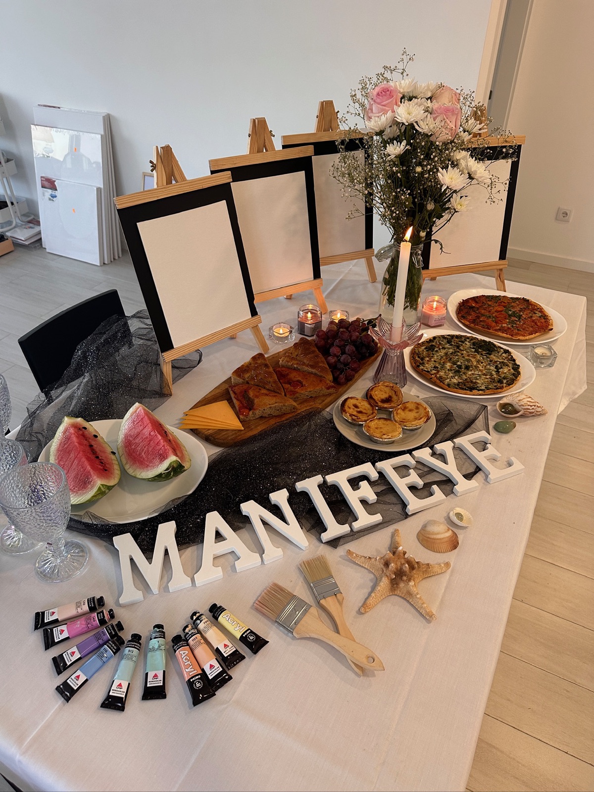 Gedeckter Workshop-Tisch mit MANIFEYE-Buchstaben, Farben, Pinseln, Leinwänden, Melone und Blumen