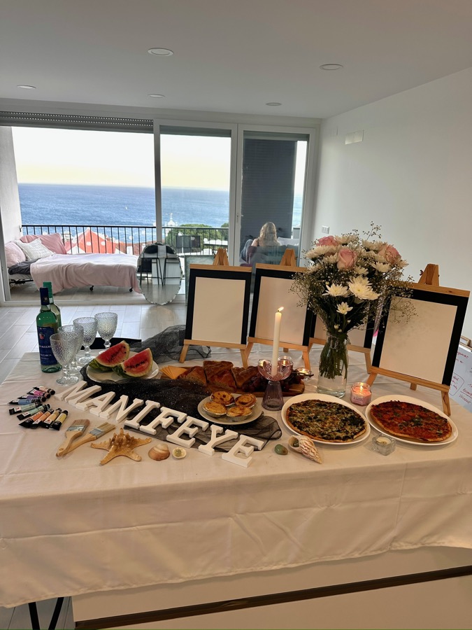 Workshop-Tisch mit Essen und Staffeleien vor großen Fenstern mit Meerblick