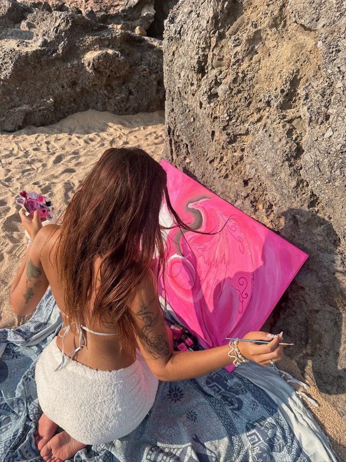 Eine Frau malt am Strand zwischen Felsen auf einer pinken Leinwand